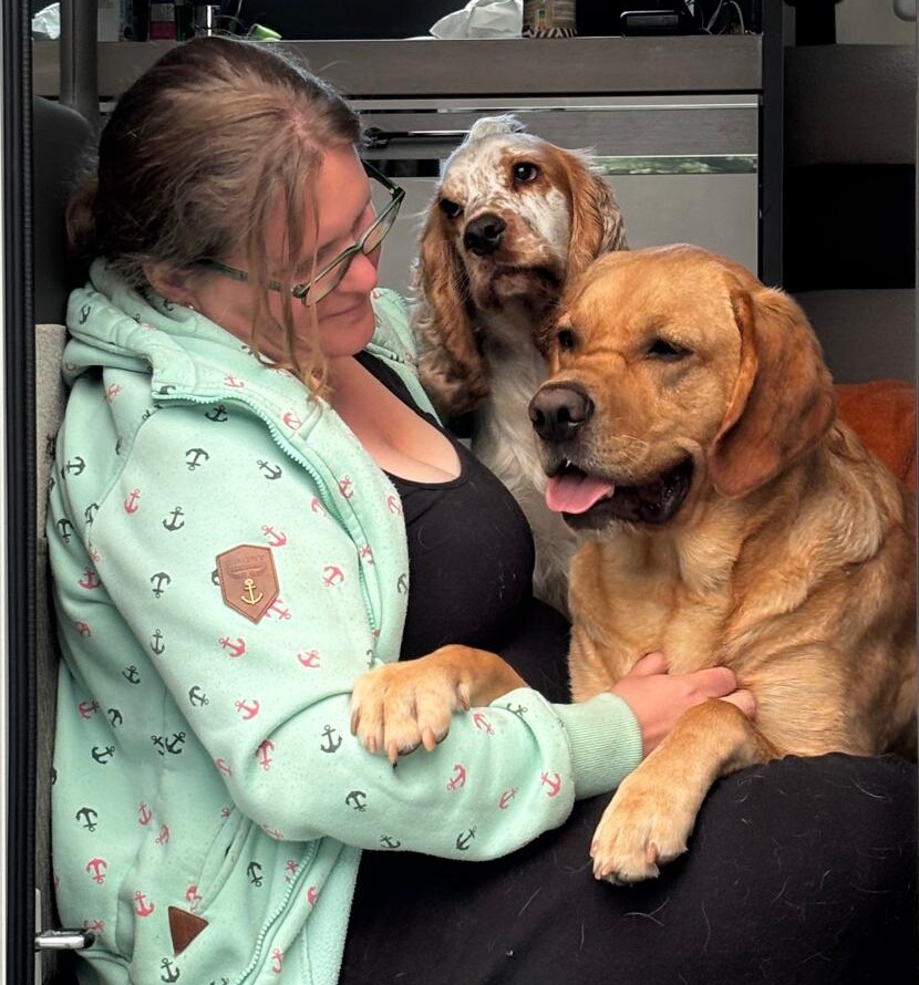 Merle mit Muffin und Fiete Ruhrpawt Blonde Frau im Profil mit einem Labrador Mischling auf dem Schoss und einem Cocker Spaniel im Hintergrund, der sie anschaut