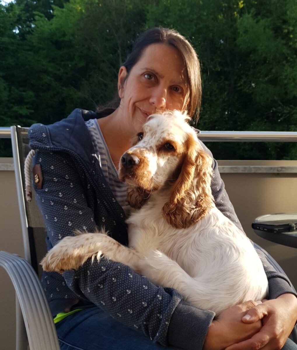 Portrait Steffi Gründerin von Ruhrpawt sitzt auf einem Stuhl auf einem Balkon mit einem roan orange Cocker Spaniel auf dem Schoß und lächelt in die Kamera